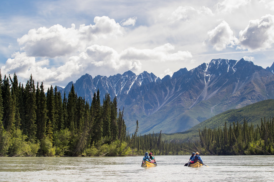 Two yellow canoes are paddling in the front of the mountains on a calm stretch of the Nahanni River, the most iconic canoe trip in Canada.