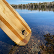 BB Special wooden canoe paddle with the tip of the blade resting in the shallow pebbles on the water
